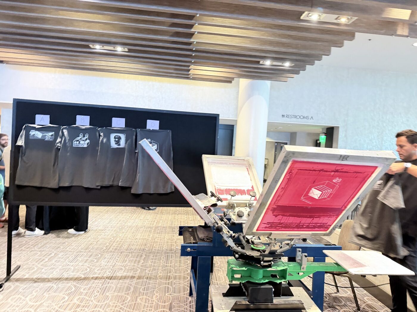 A daylit live-printing booth with a loaded press and finished merch stacked on the tables