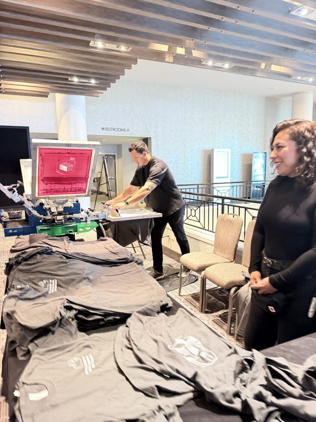 A printer working the press at a bright daytime event beside stacks of folded tees